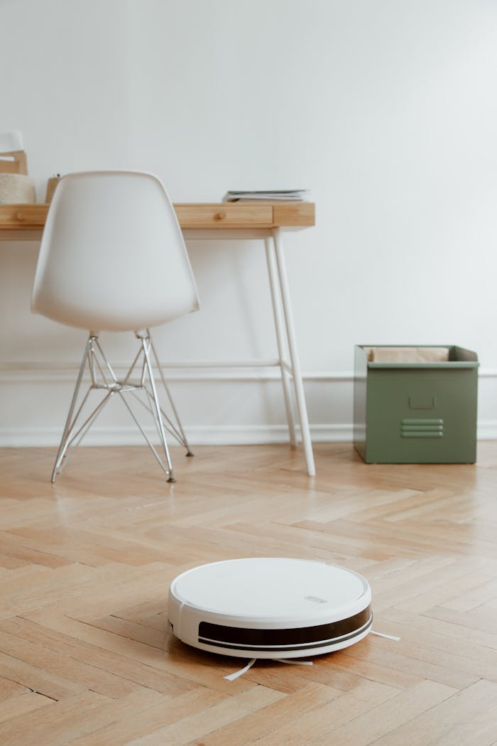 A modern minimalist room featuring a robotic vacuum cleaner on a wooden floor beside a desk and chair.