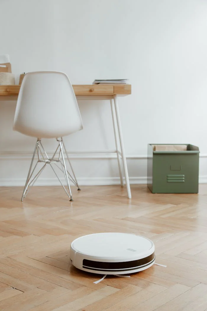 A modern minimalist room featuring a robotic vacuum cleaner on a wooden floor beside a desk and chair.